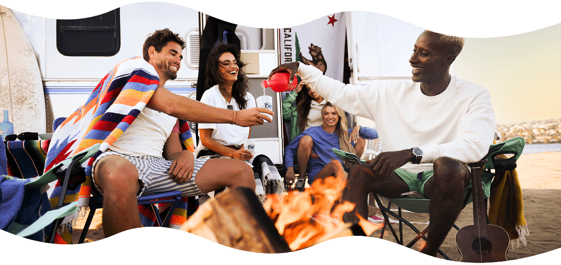 A multiracial group of friends surrounding a campfire share a bottle of Beach Whiskey.